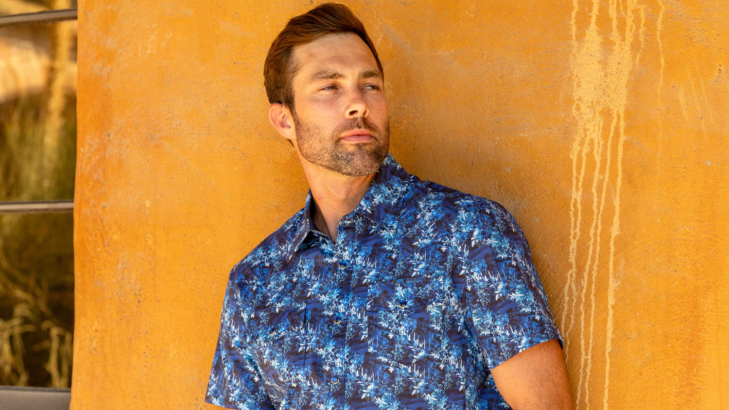 A close-up of a man wearing a blue short-sleeve button-down shirt with a vibrant abstract geometric pattern, leaning against a textured orange wall.