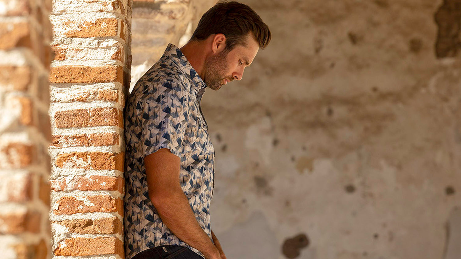 A side-profile shot of a man wearing a blue short-sleeve button-down shirt with a multi-shaded abstract geometric print, looking down while leaning against a textured neutral tan wall.