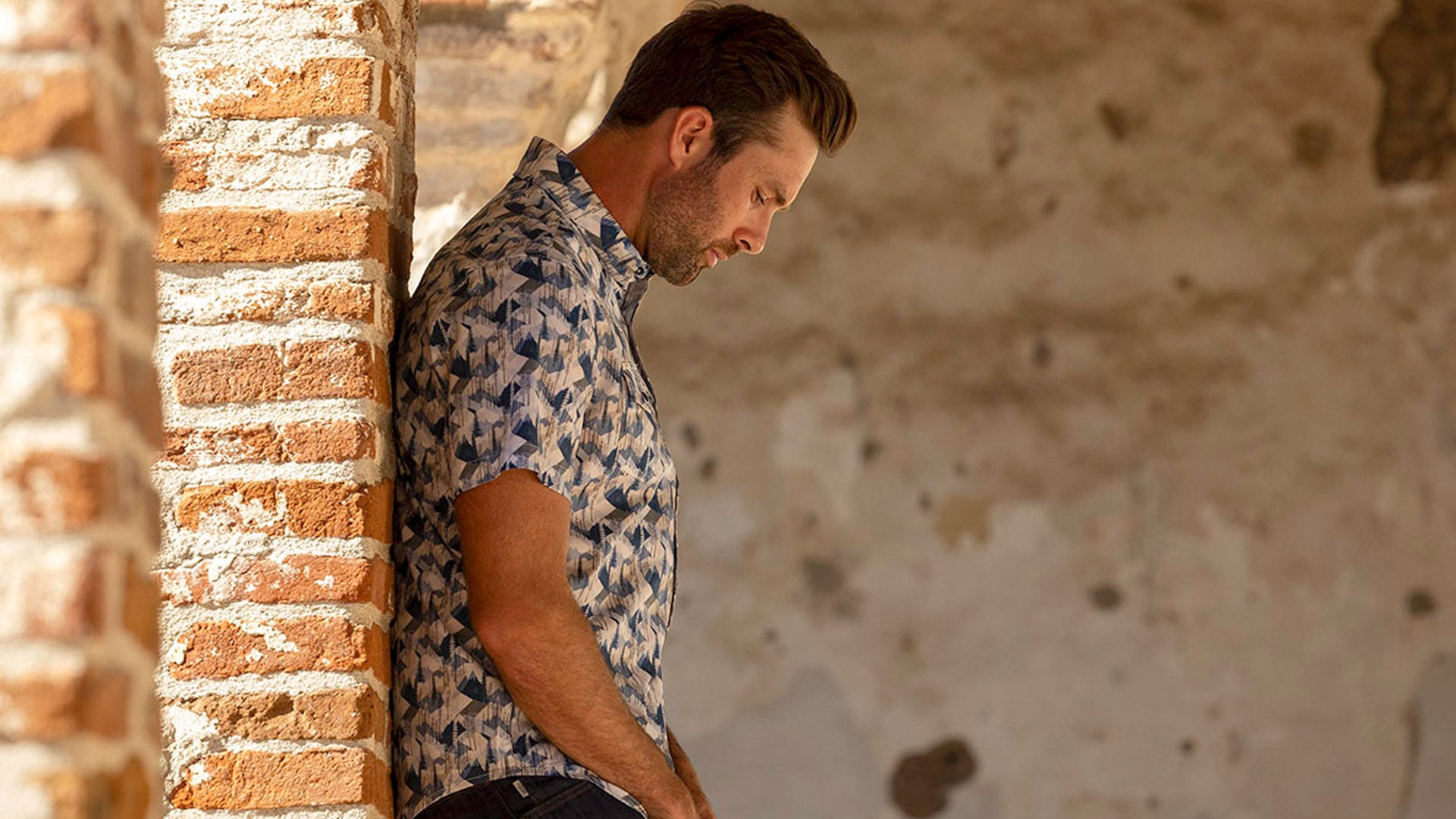 A side-profile shot of a man wearing a blue short-sleeve button-down shirt with a multi-shaded abstract geometric print, looking down while leaning against a textured neutral tan wall.
