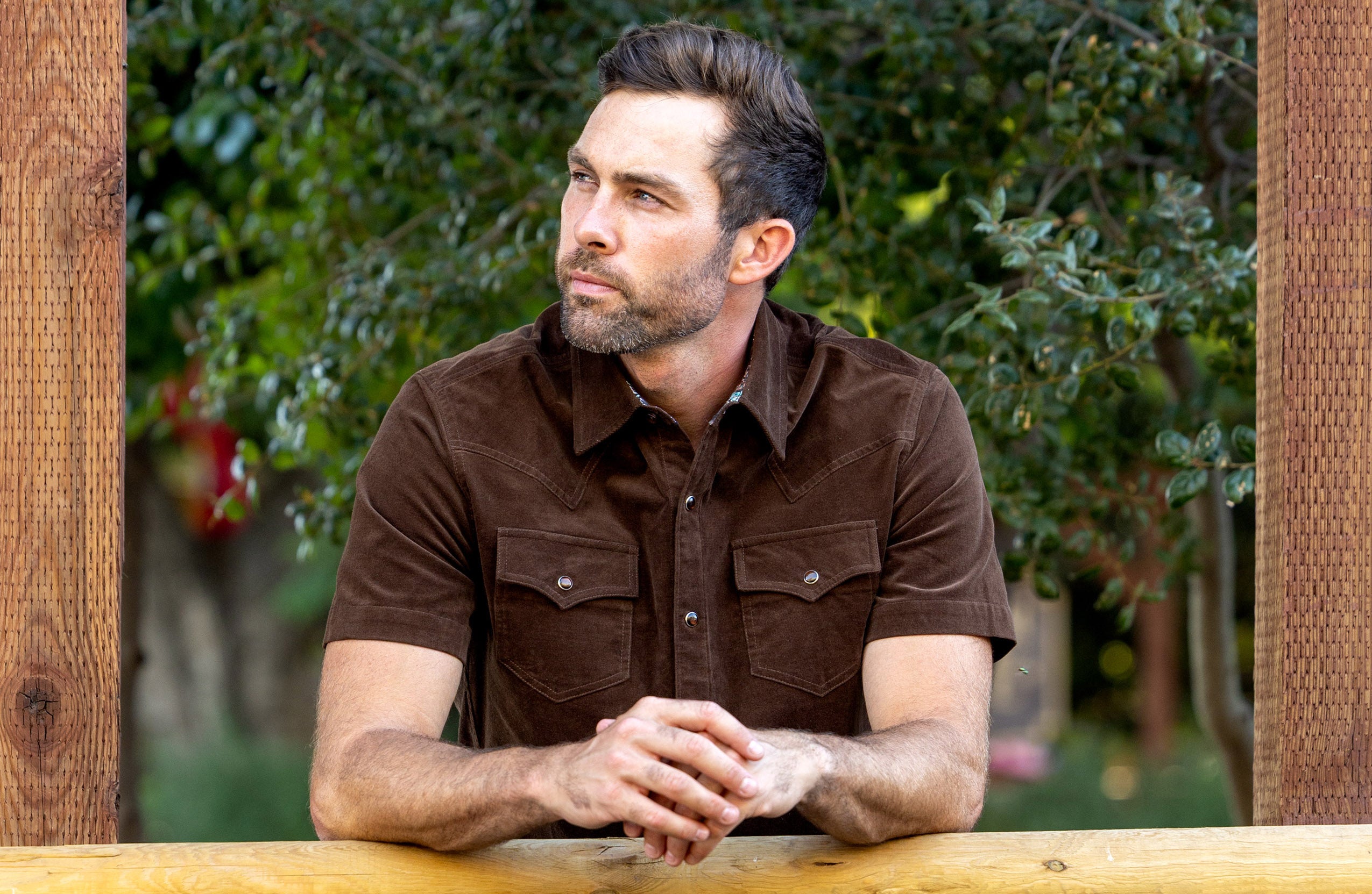 A man sits outdoors at a wooden table wearing a brown fine-wale corduroy short-sleeve button-down shirt with metallic snap buttons and western chest pockets.