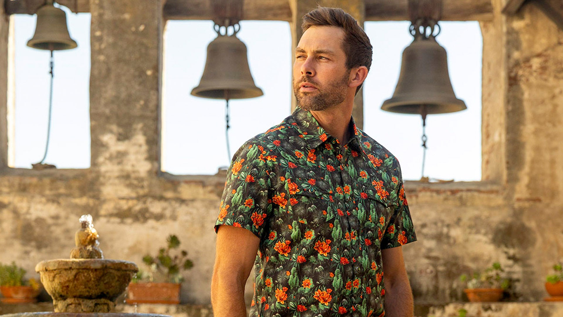 A man stands outdoors in front of a stone wall archway featuring hanging metal bells, wearing a black short-sleeve button-down western shirt with a vibrant orange floral and green cactus pattern.