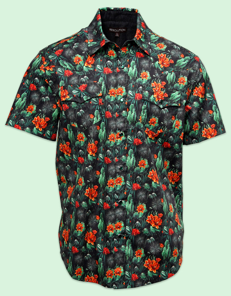 A black short-sleeve button-down western shirt featuring a vibrant orange floral and green cactus pattern, detailed with double western-style chest pockets against a light gray background.