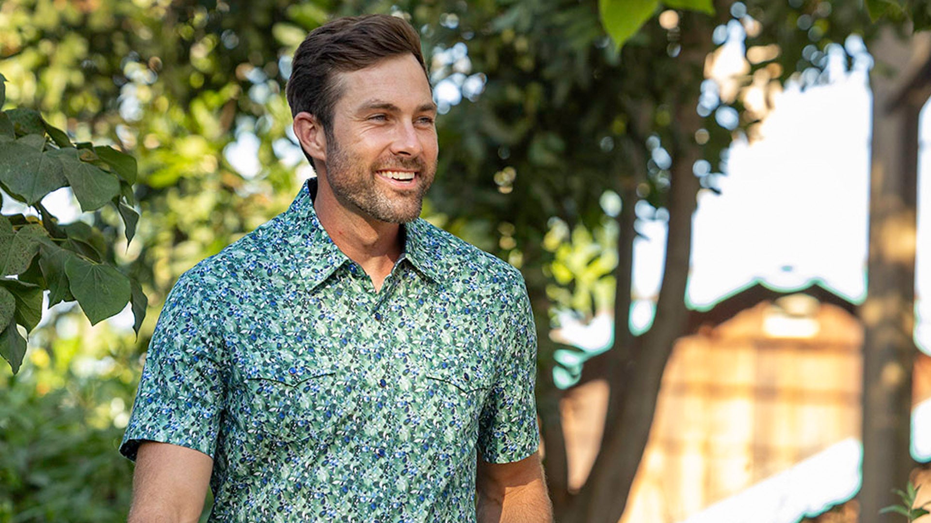 A smiling man stands outdoors with blurred green trees in the background, wearing a green short-sleeve button-down shirt featuring a dense black and white houndstooth pattern. This premium performance top offers a tailored fit designed for active movement and elevated everyday style.